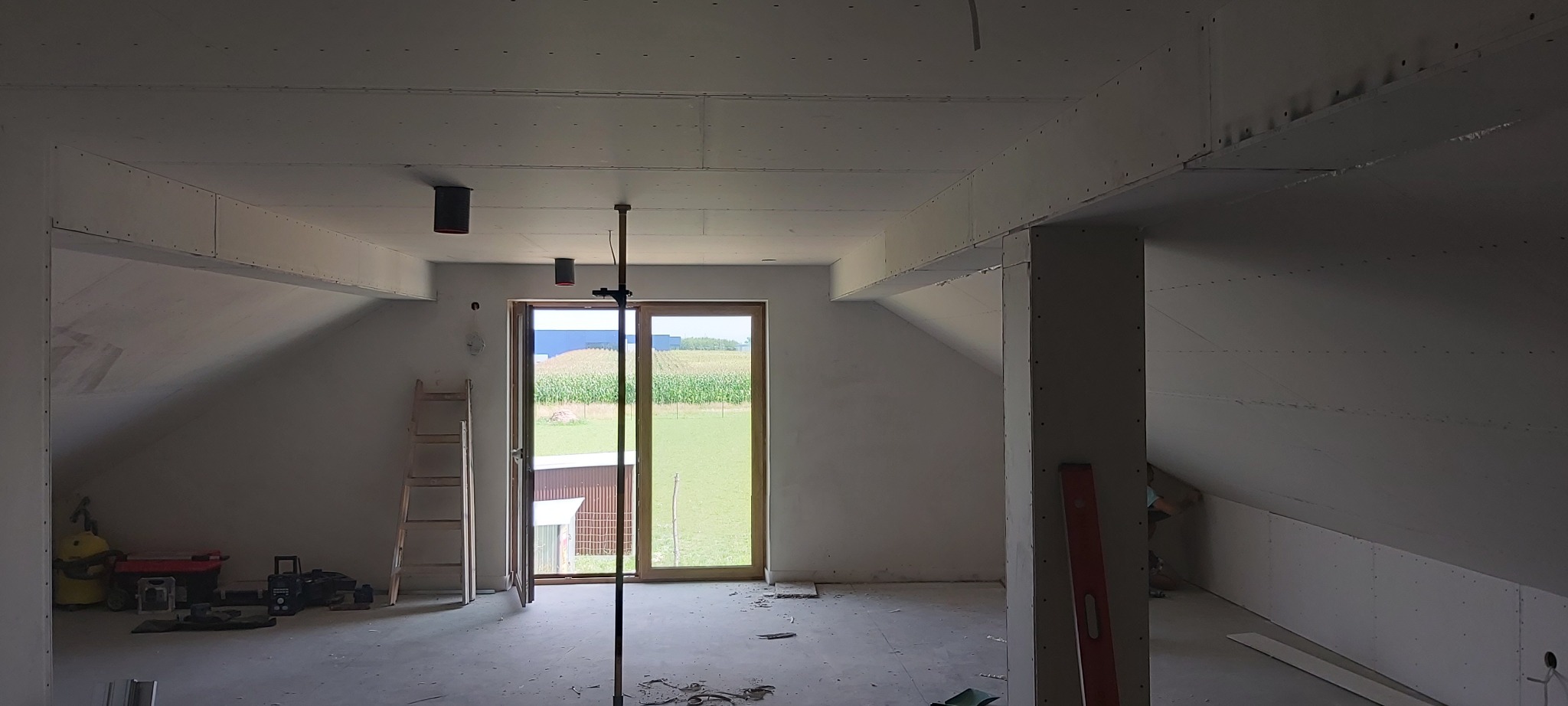 Poddasze w trakcie prac wykończeniowych: widoczne płyty kartonowo-gipsowe na ścianach i suficie, drabina oparta o ścianę, narzędzia budowlane w rogu oraz widok na pole kukurydzy przez otwarte drzwi...