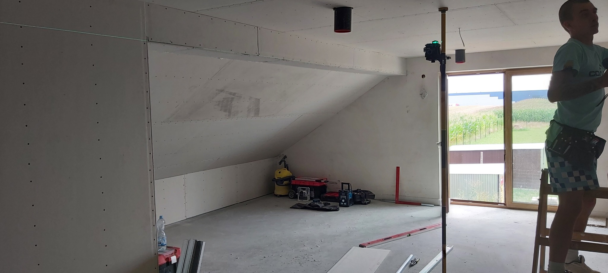 Montaż płyt kartonowo-gipsowych na poddaszu w Wodzisławiu Śląskim. Widoczny pracownik na drabinie, laser krzyżowy na statywie, przymocowane płyty z widocznymi wkrętami oraz narzędzia i odkurzacz...