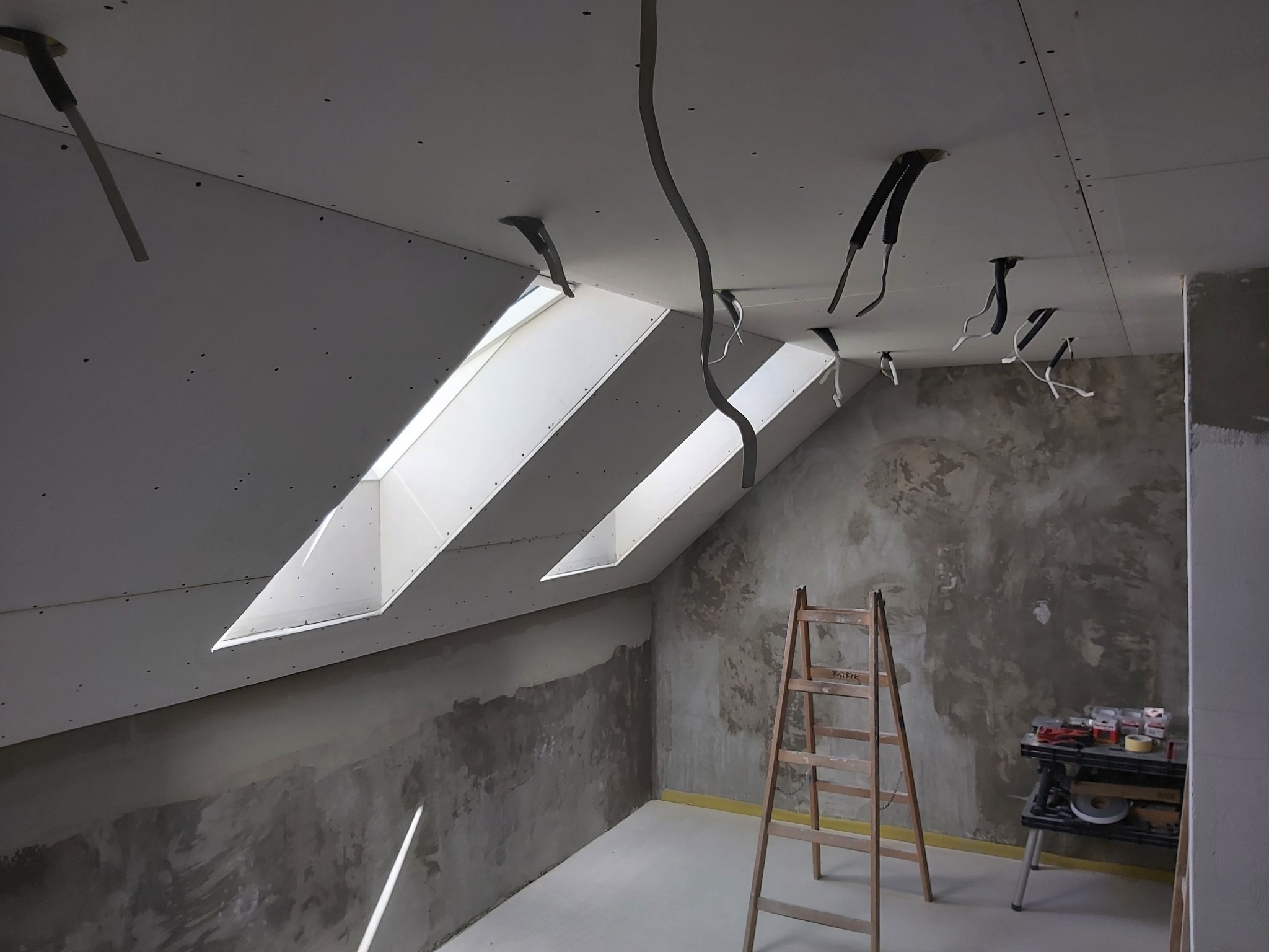 Poddasze w trakcie prac wykończeniowych: zamontowane płyty karton-gips, widoczne kable elektryczne, okna dachowe, drabina oparta o ścianę z surowym wykończeniem, narzędzia na stole roboczym.