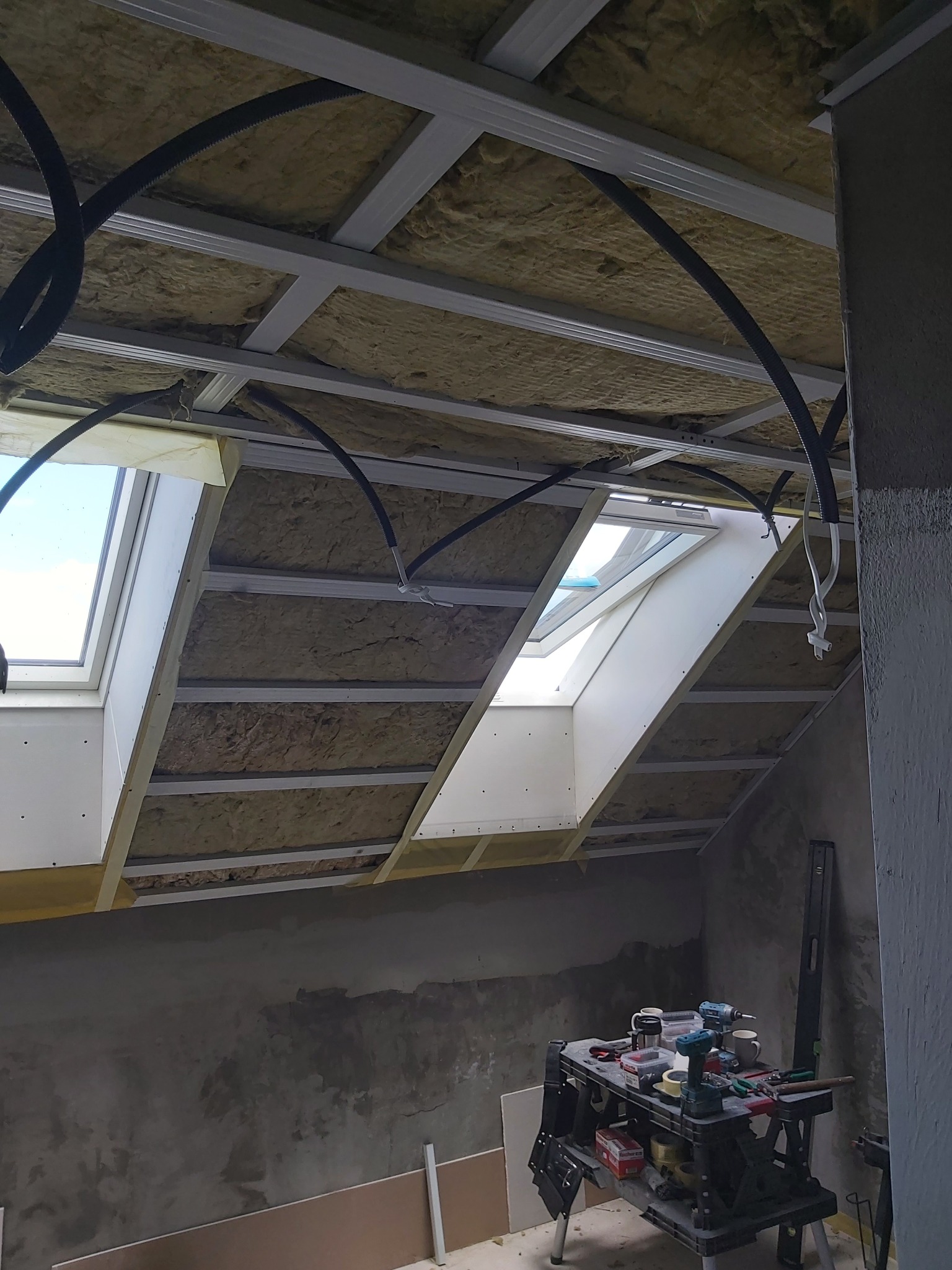 Poddasze w trakcie adaptacji: widoczne okna dachowe, ocieplenie wełną mineralną między krokwiami, profile metalowe, instalacja elektryczna w peszlach oraz stół roboczy z narzędziami i poziomica.