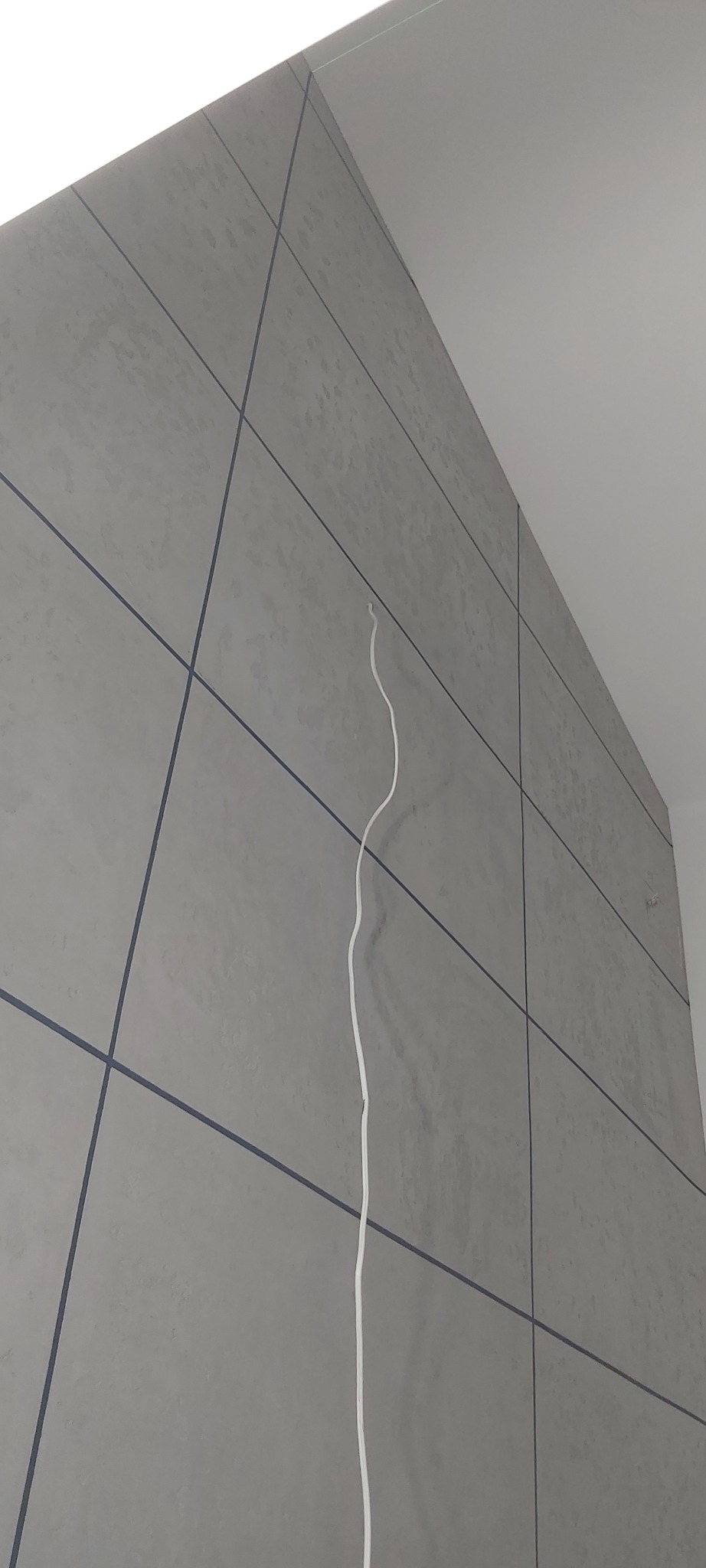 Szare płytki ścienne z ciemnymi fugami i wystającą białą listwą przypodłogową, widok z dołu.