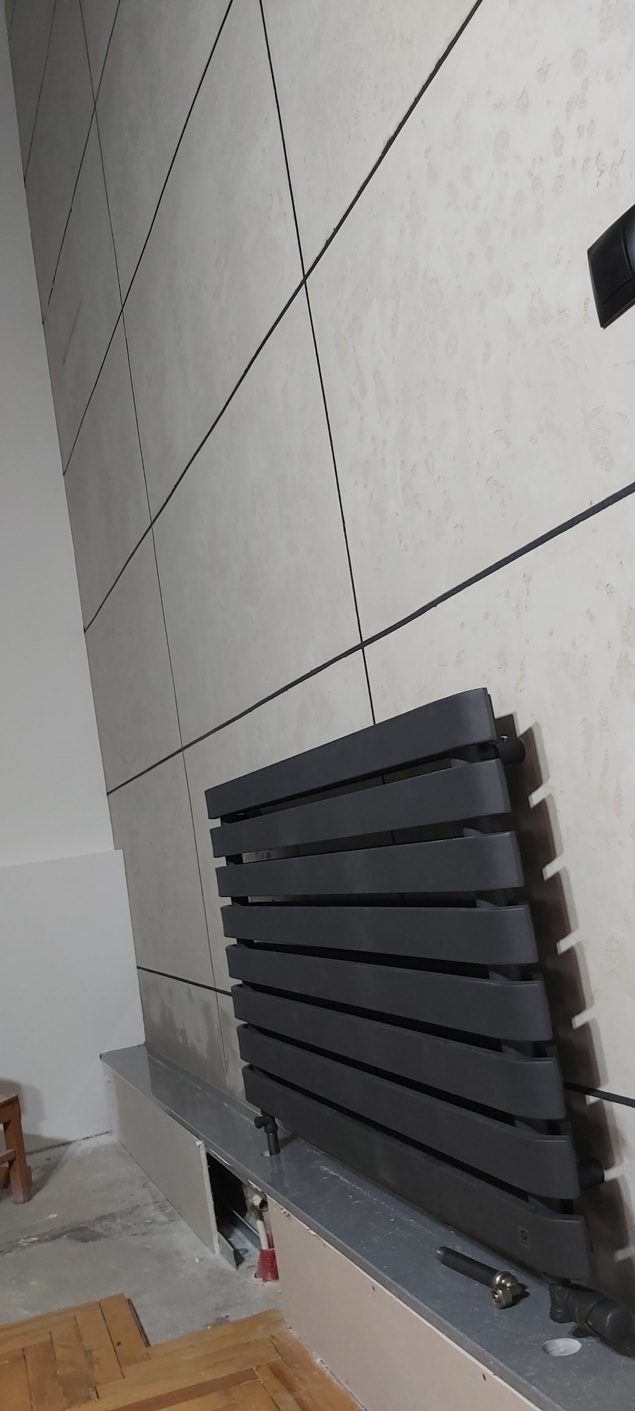 Czarny grzejnik dekoracyjny zamontowany na ścianie wyłożonej jasnymi, kwadratowymi płytkami z ciemnymi fugami, widoczne podłączenia i fragment podłogi w trakcie prac wykończeniowych.