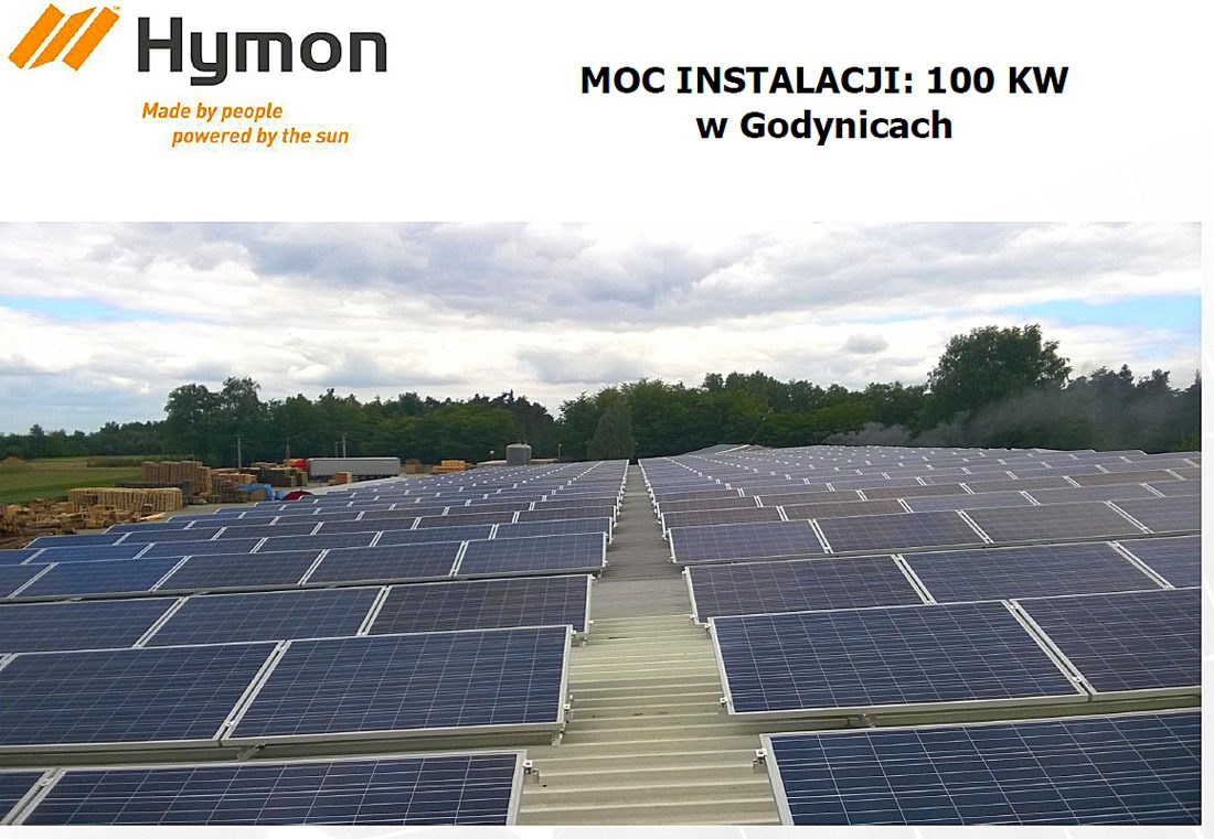 Rozległa instalacja paneli słonecznych na dachu budynku, widok z góry, w tle drzewa i pochmurne niebo, w lewym górnym rogu logo firmy.