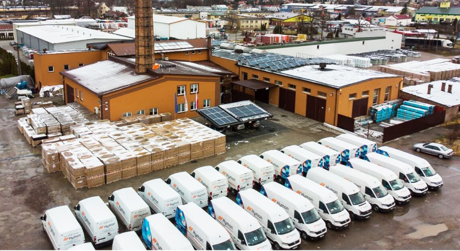 Widok z góry na teren firmy z zaparkowanymi samochodami dostawczymi z logo, paletami materiałów budowlanych, budynkami z panelami słonecznymi na dachach oraz wolnostojącym systemem paneli na ziemi.