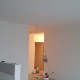 Wnętrze w trakcie malowania: białe ściany, sufit, widoczne puszki z farbą, narzędzia malarskie i wisząca żarówka