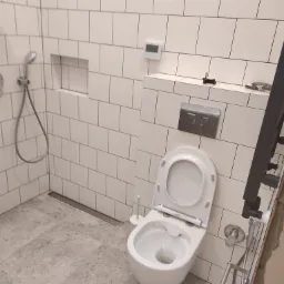 Nowoczesna toaleta z otwartą klapą w łazience wykończonej białymi, prostokątnymi płytkami i szarą podłogą imitującą kamień, widoczny odpływ liniowy w podłodze.