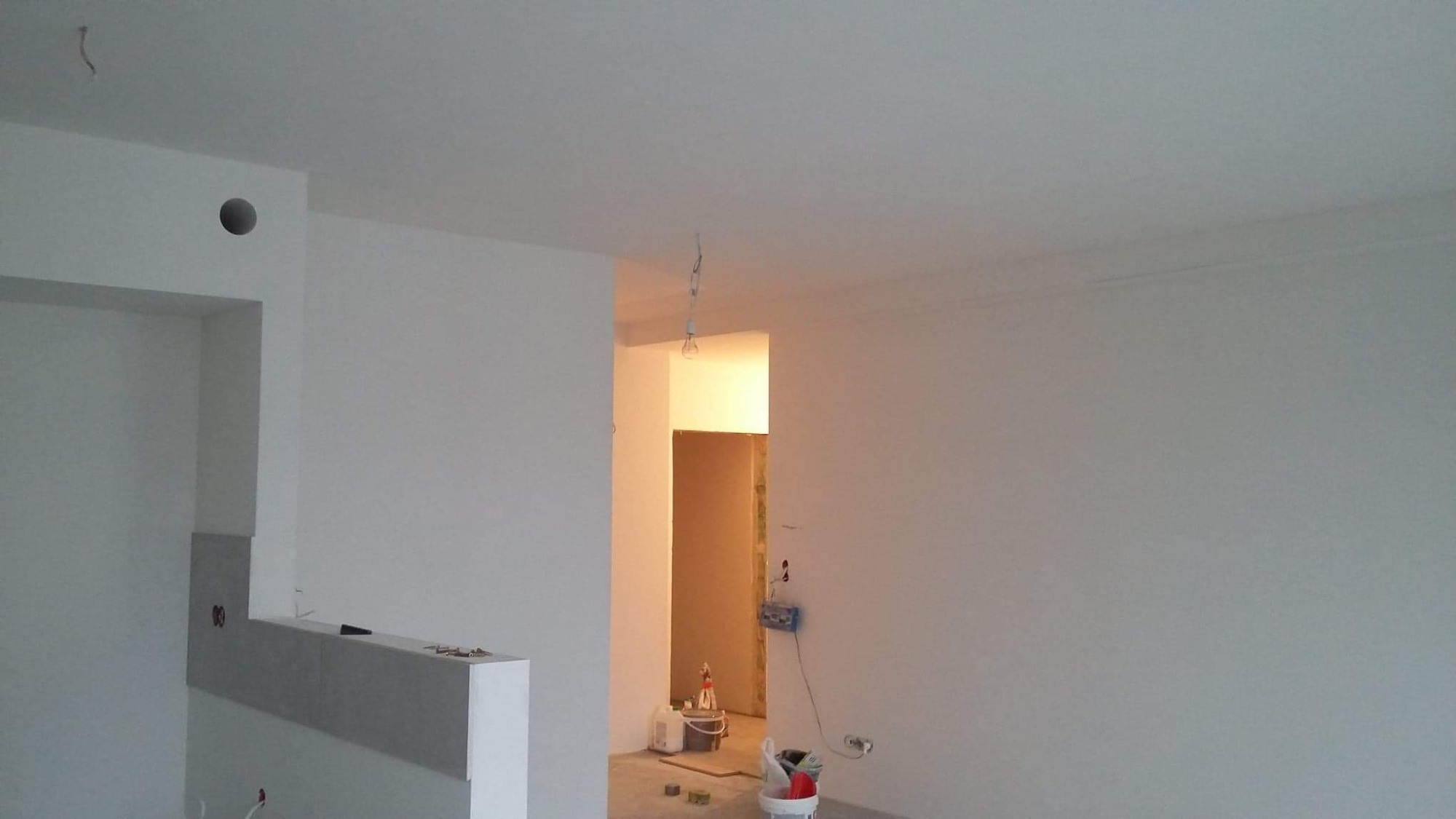 Wnętrze w trakcie malowania: białe ściany, sufit, widoczne puszki z farbą, narzędzia malarskie i wisząca żarówka