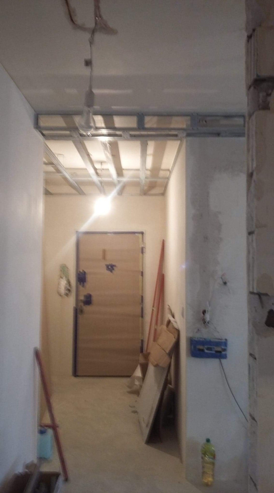 Korytarz w trakcie prac wykończeniowych: widoczna konstrukcja sufitu podwieszanego z profili metalowych, surowe ściany, drzwi wejściowe i materiały budowlane.