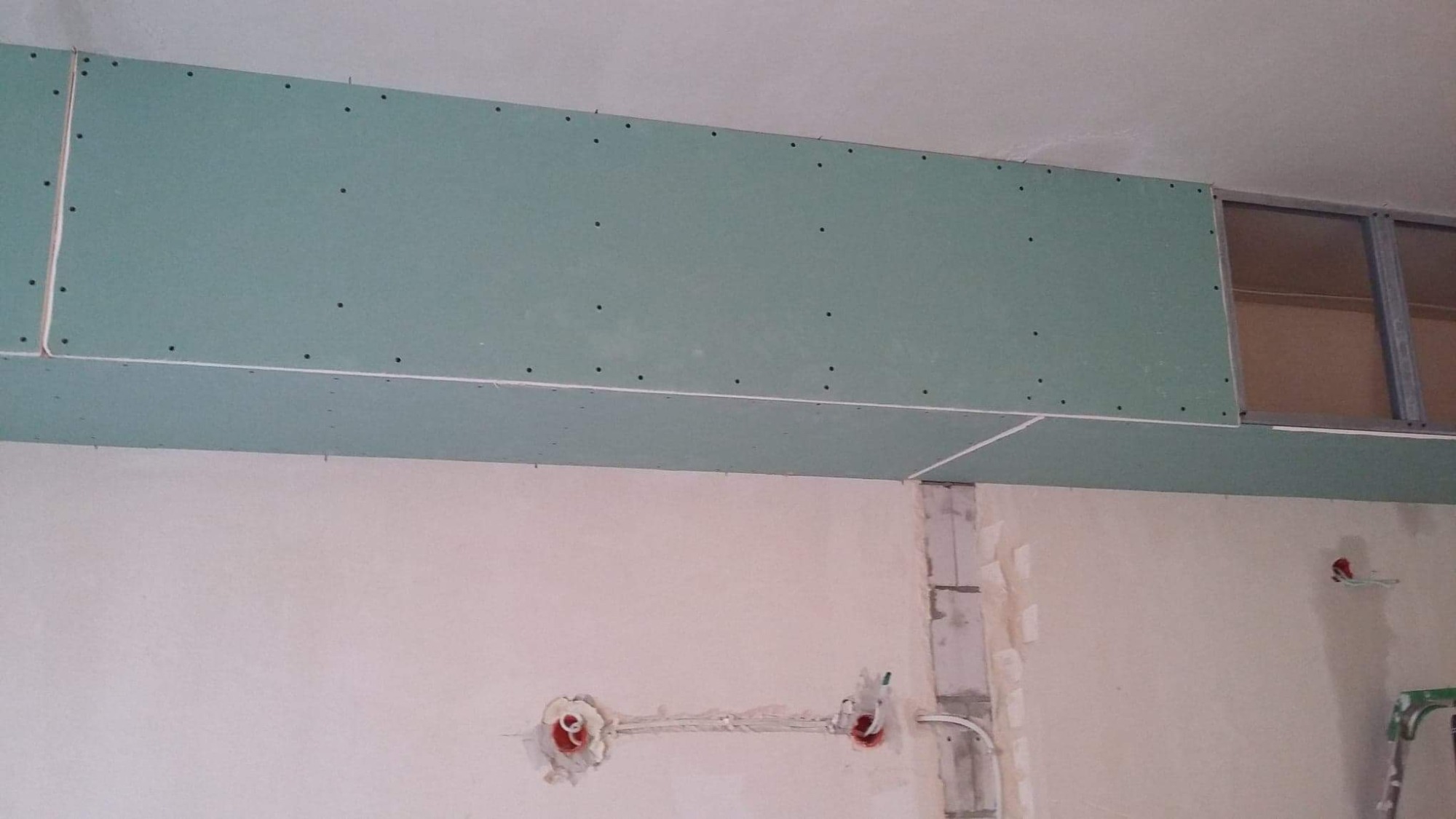 Montaż zielonych płyt gipsowo-kartonowych na suficie, widoczne metalowe profile, przygotowane otwory na instalacje elektryczne w ścianie.