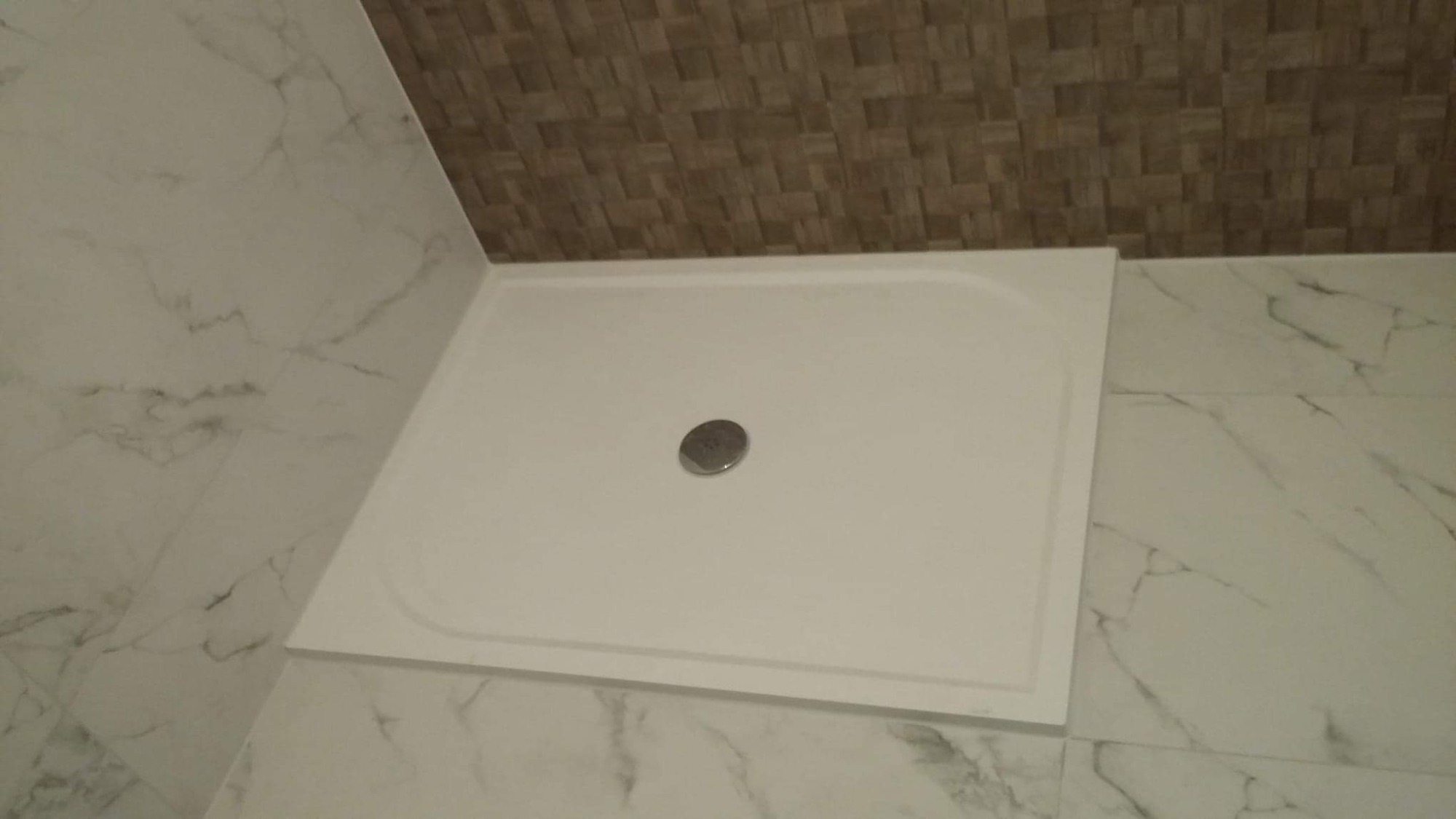 Biały brodzik z odpływem w rogu łazienki wyłożonej marmurowymi płytkami i mozaiką na suficie.