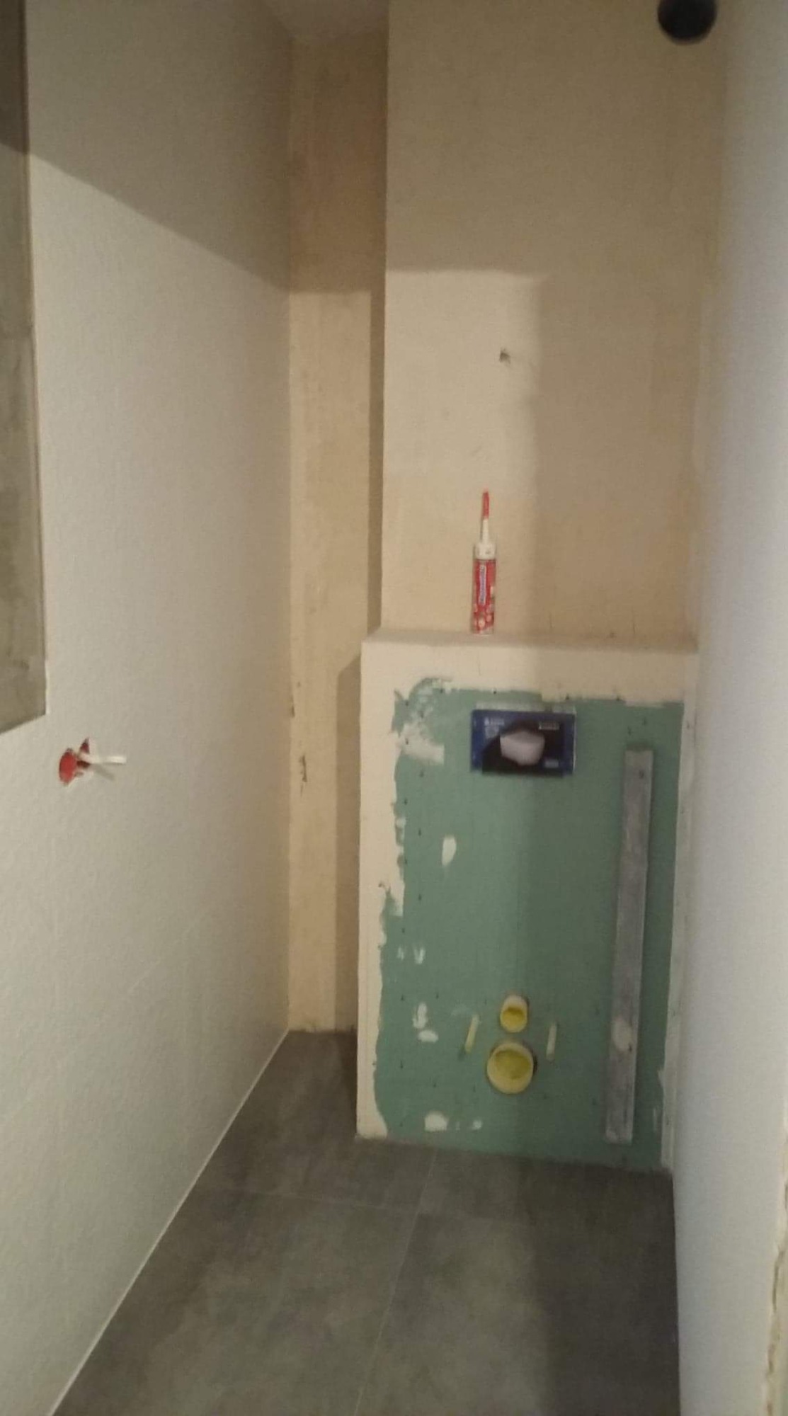 Wnętrze łazienki w trakcie prac: widoczne szare płytki podłogowe, częściowo przygotowane ściany z płyt gipsowo-kartonowych, elementy instalacji wodnej oraz pionowa listwa pomiarowa przy ścianie...