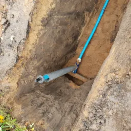 Głęboki wykop w ziemi z widocznym niebieskim rurociągiem oraz szarą rurą z czarnym kablem w środku, połączonymi z istniejącą instalacją, ziemia brudna, ślady opon, fragment buta w prawym dolnym rogu.