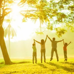 Rodzina skacząca na zielonej łące w słoneczny dzień, pod rozłożystym drzewem, z promieniami słońca przebijającymi przez liście.