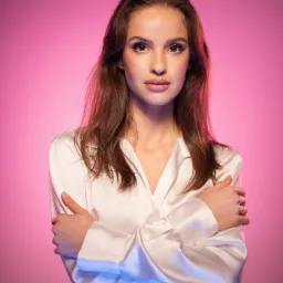 Portret kobiety w białej satynowej koszuli z rękawami skrzyżowanymi na piersiach, z delikatnym makijażem i pierścionkiem na palcu, na różowo-niebieskim tle.