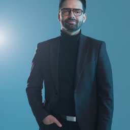 Justyna Szczygielska - Portret mężczyzny w okularach, czarnym golfie i grafitowej marynarce na jednolitym, błękitnym tle, z ręką w kieszeni i srebrnym paskiem