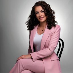 Justyna Szczygielska - Portret kobiety w różowym garniturze siedzącej na czarnym krześle na szarym tle. Uśmiechnięta brunetka z kręconymi włosami, subtelny makijaż, delikatna biżuteria.