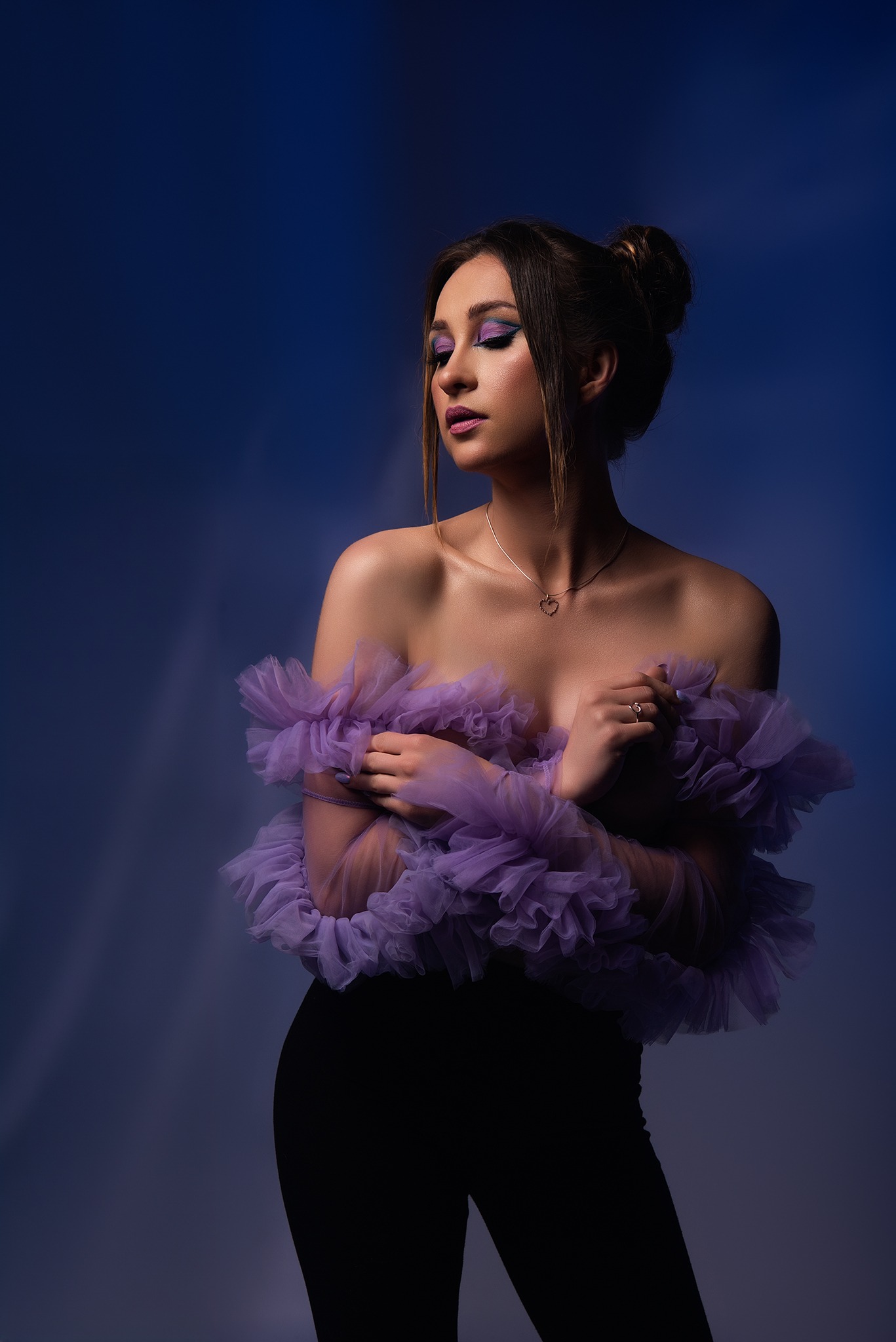Portret kobiety z zamkniętymi oczami, w czarnych spodniach i fioletowej, tiulowej kreacji, na ciemnoniebieskim tle z subtelnym oświetleniem.