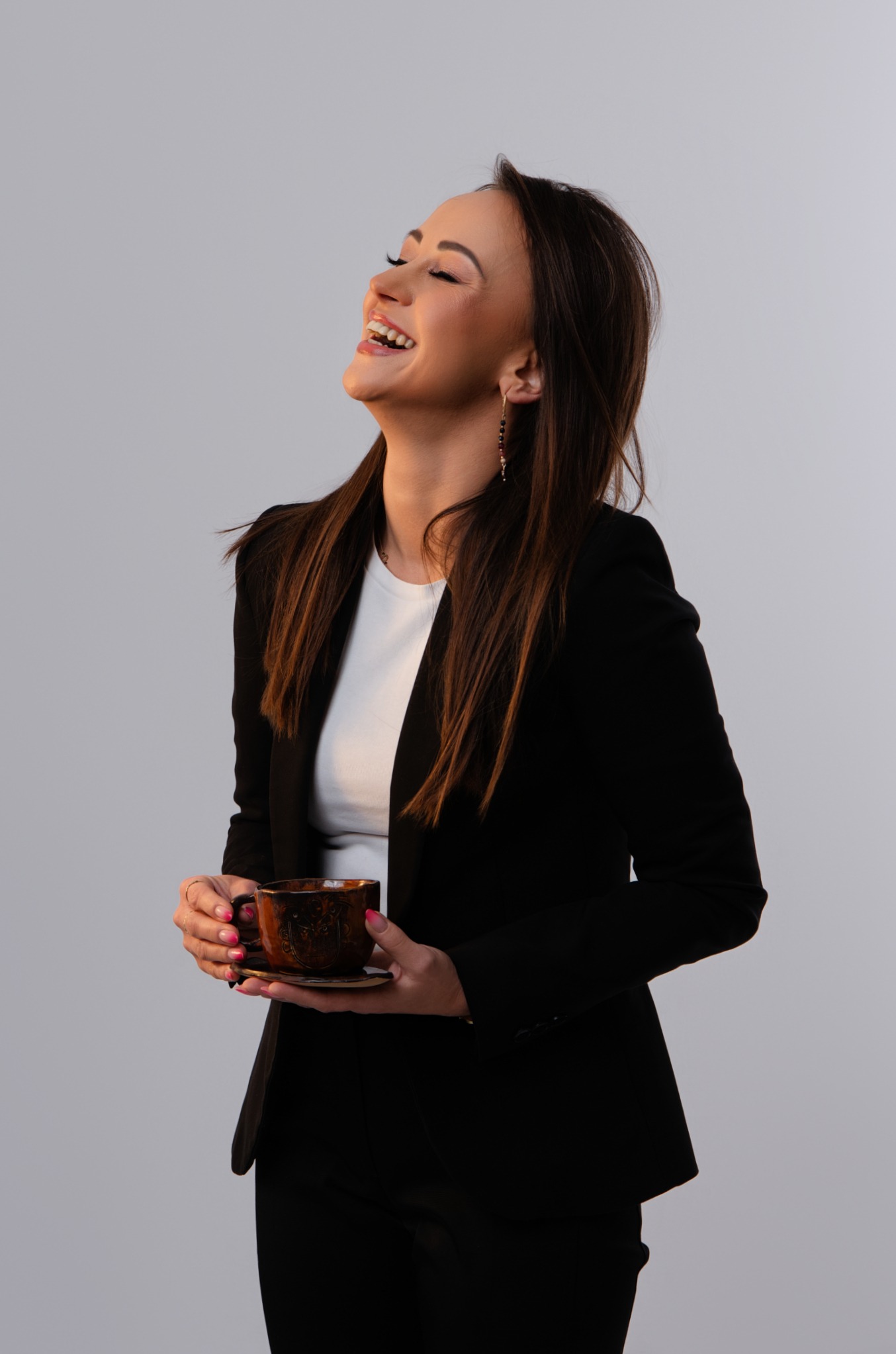 Uśmiechnięta brunetka w czarnej marynarce i białej koszulce, trzymająca zdobioną filiżankę z kawą, głowa odchylona do tyłu, widoczne kolczyki i delikatny makijaż.