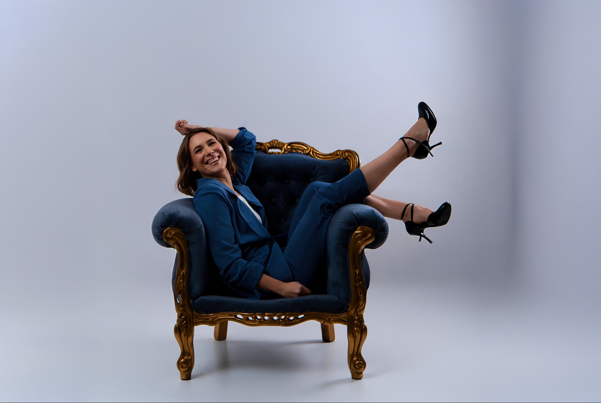 Uśmiechnięta kobieta w niebieskim garniturze relaksuje się w aksamitnym fotelu z pozłacanymi zdobieniami, eksponując czarne szpilki na tle jasnego studia.