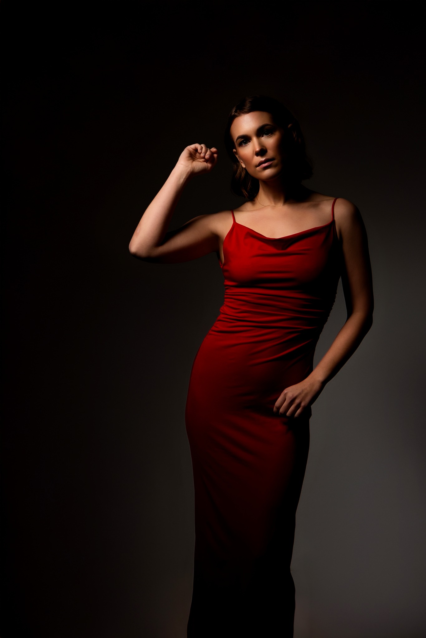 Kobieta w czerwonej, drapowanej sukience na ramiączkach, pozująca na ciemnym tle, z ręką uniesioną do góry i spojrzeniem skierowanym w dół.