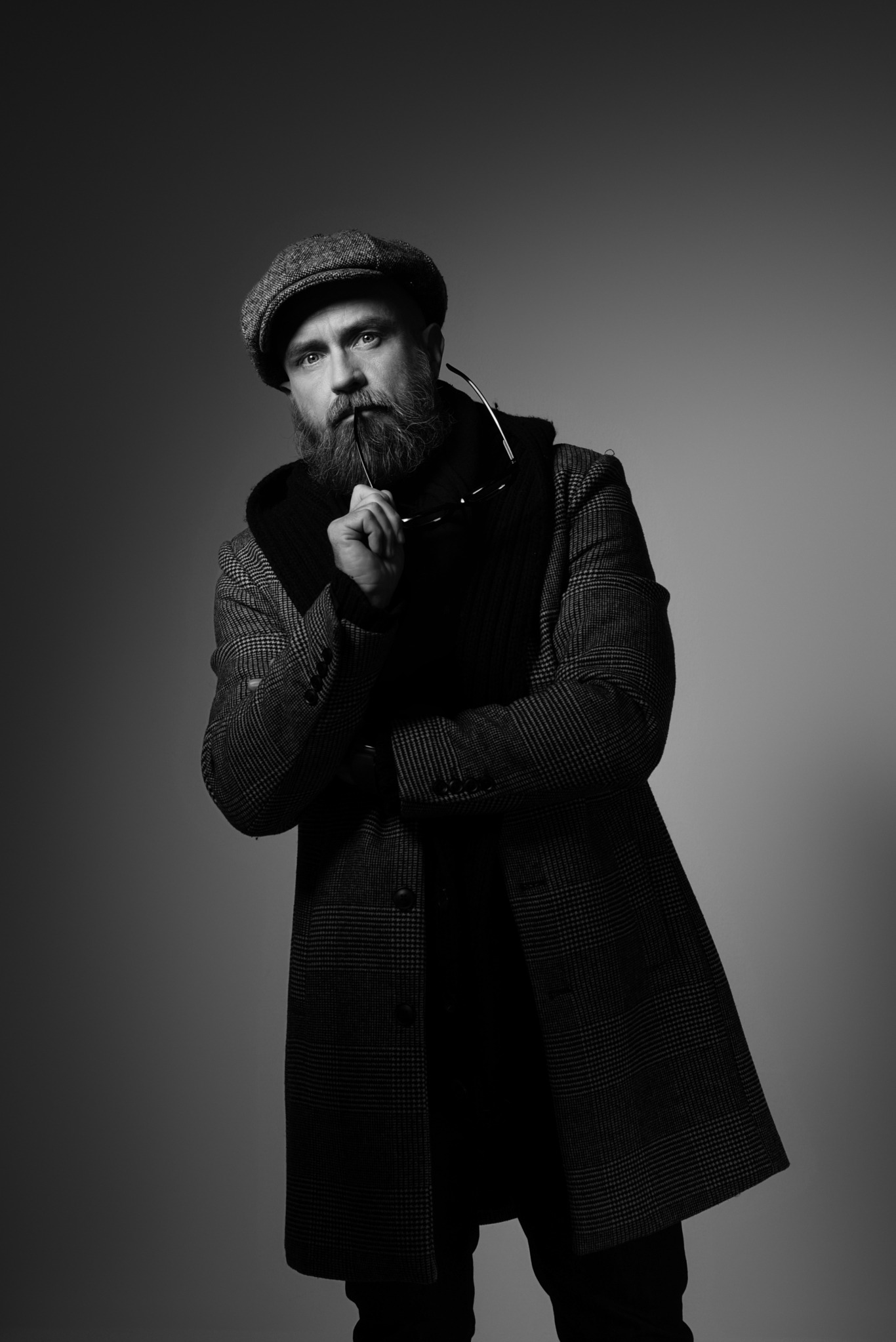 Czarno-biały portret mężczyzny z brodą, w kaszkiecie i płaszczu w kratę, trzymającego okulary przy ustach, na jednolitym szarym tle.