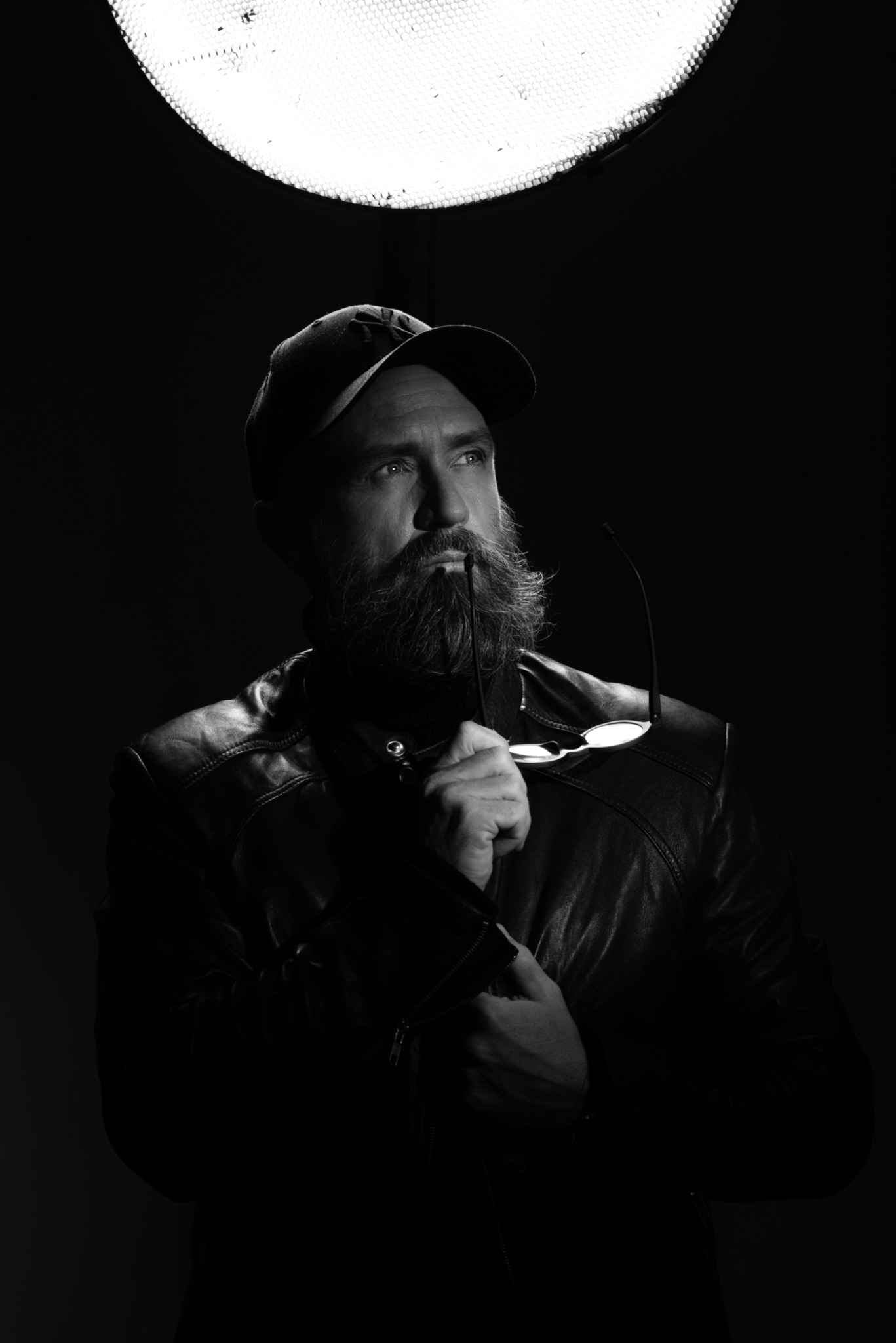 Czarno-biały portret mężczyzny z brodą w skórzanej kurtce i czapce, trzymającego okulary przy ustach, oświetlonego od góry lampą studyjną, zamyślony wzrok skierowany w górę.