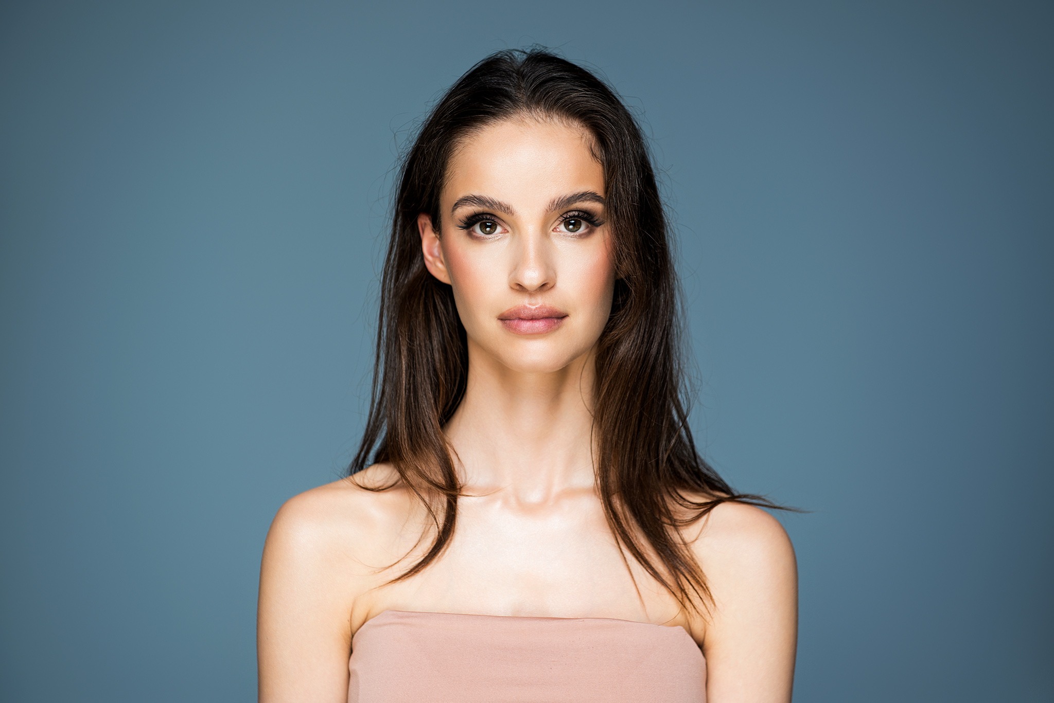 Portret młodej kobiety z długimi brązowymi włosami, delikatnym makijażem i beżową tkaniną owiniętą wokół ramion, na jednolitym niebieskim tle.
