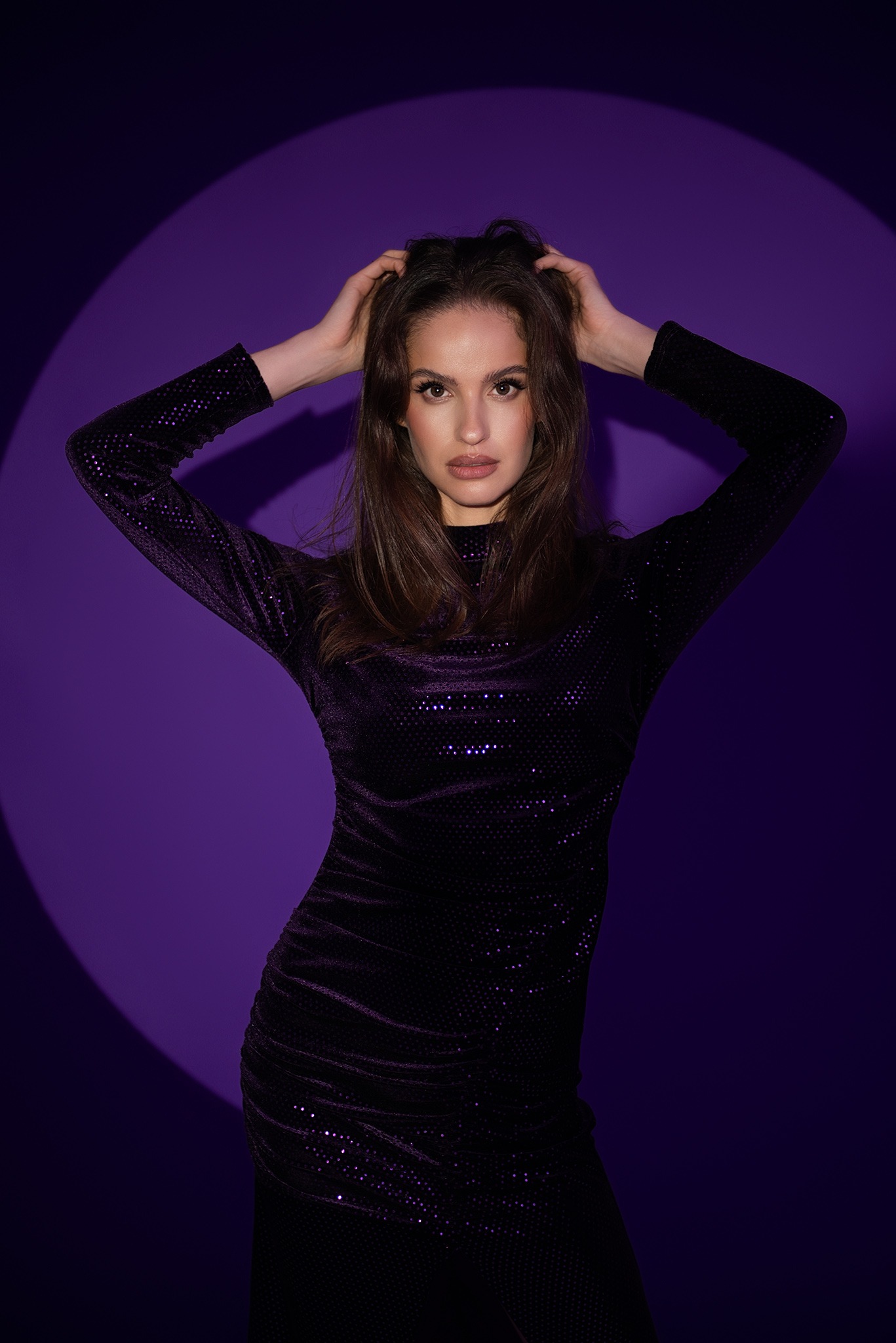 Portret kobiety w czarnej, błyszczącej sukience na fioletowym tle oświetlonym punktowym światłem, dłonie uniesione do włosów, spojrzenie skierowane w obiektyw.