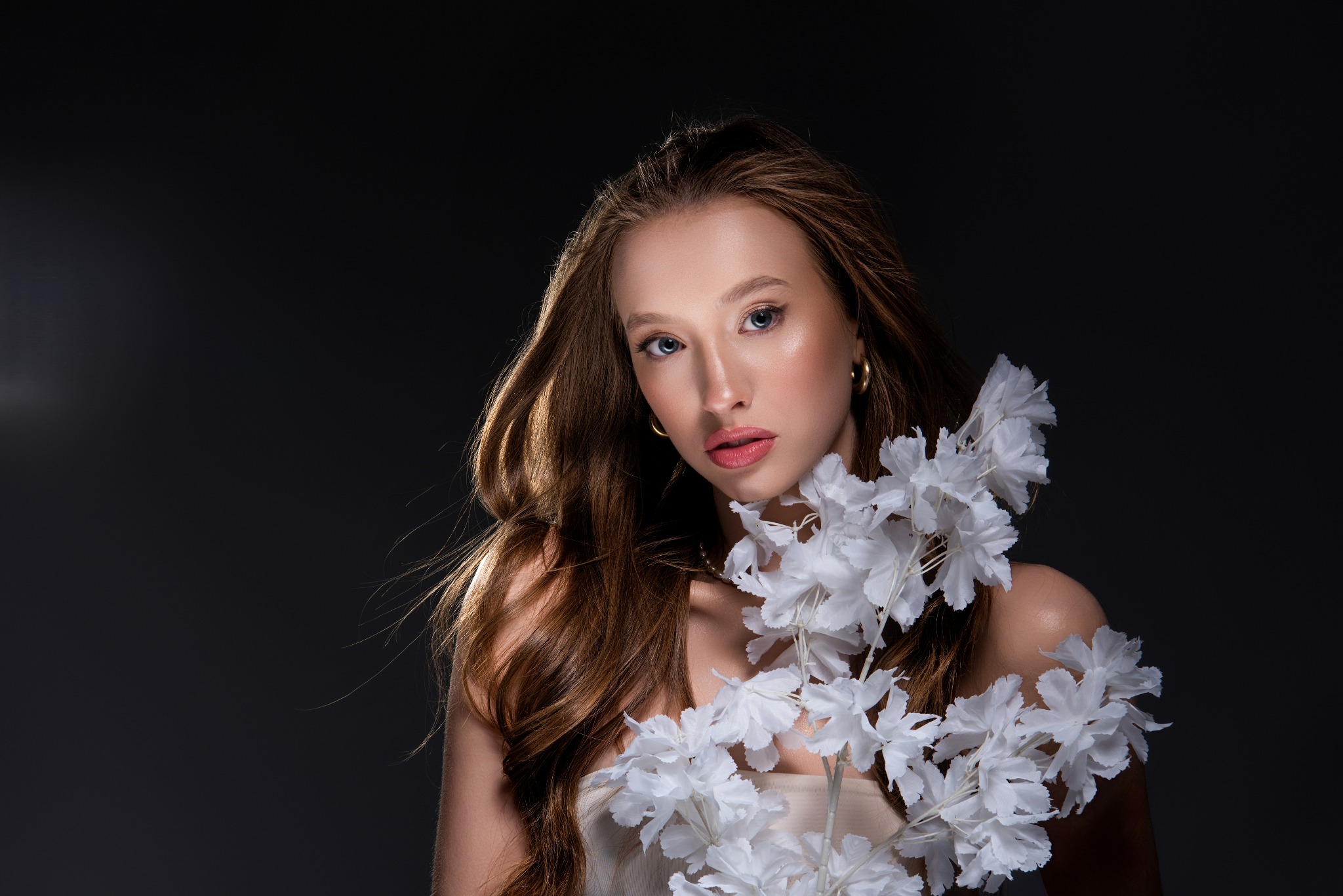 Portret kobiety z długimi, brązowymi włosami i białymi, papierowymi kwiatami na ramieniu, na ciemnoszarym tle. Modelka ma delikatny makijaż i złote kolczyki.