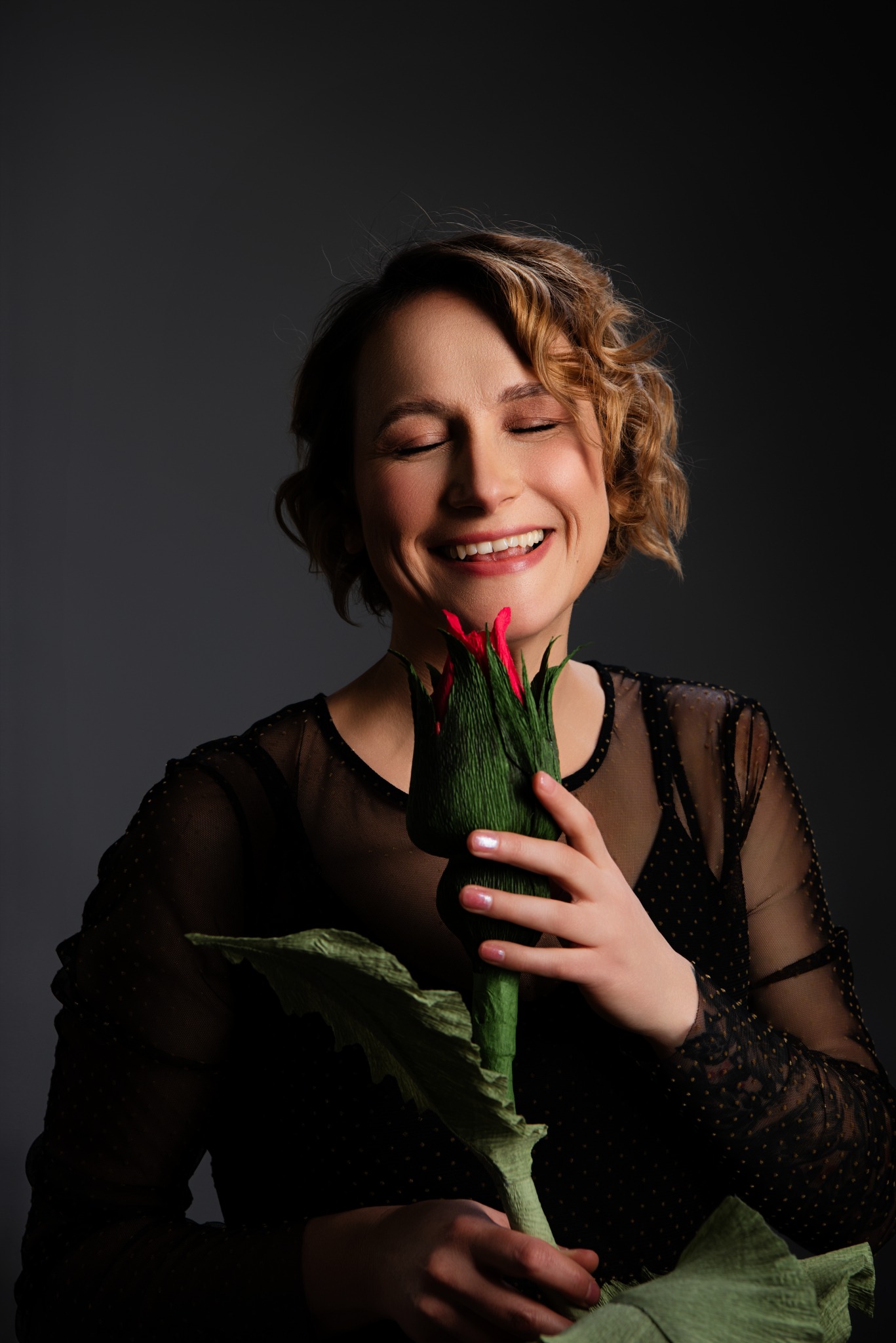 Uśmiechnięta kobieta z zamkniętymi oczami trzyma duży, stylizowany kwiat z papieru w odcieniach zieleni i czerwieni na ciemnoszarym tle.