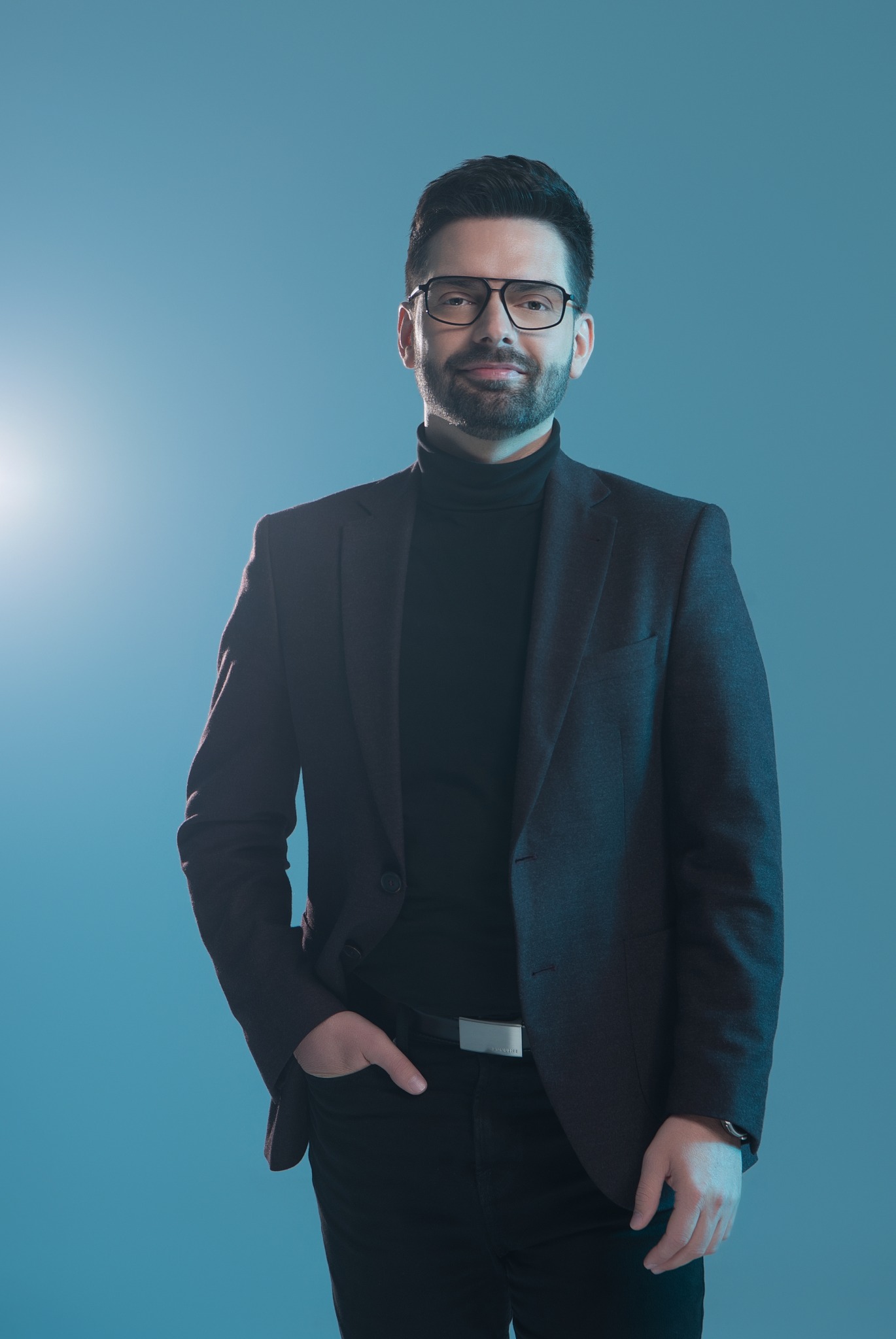 Portret mężczyzny w okularach, czarnym golfie i grafitowej marynarce na jednolitym, błękitnym tle, z ręką w kieszeni i srebrnym paskiem