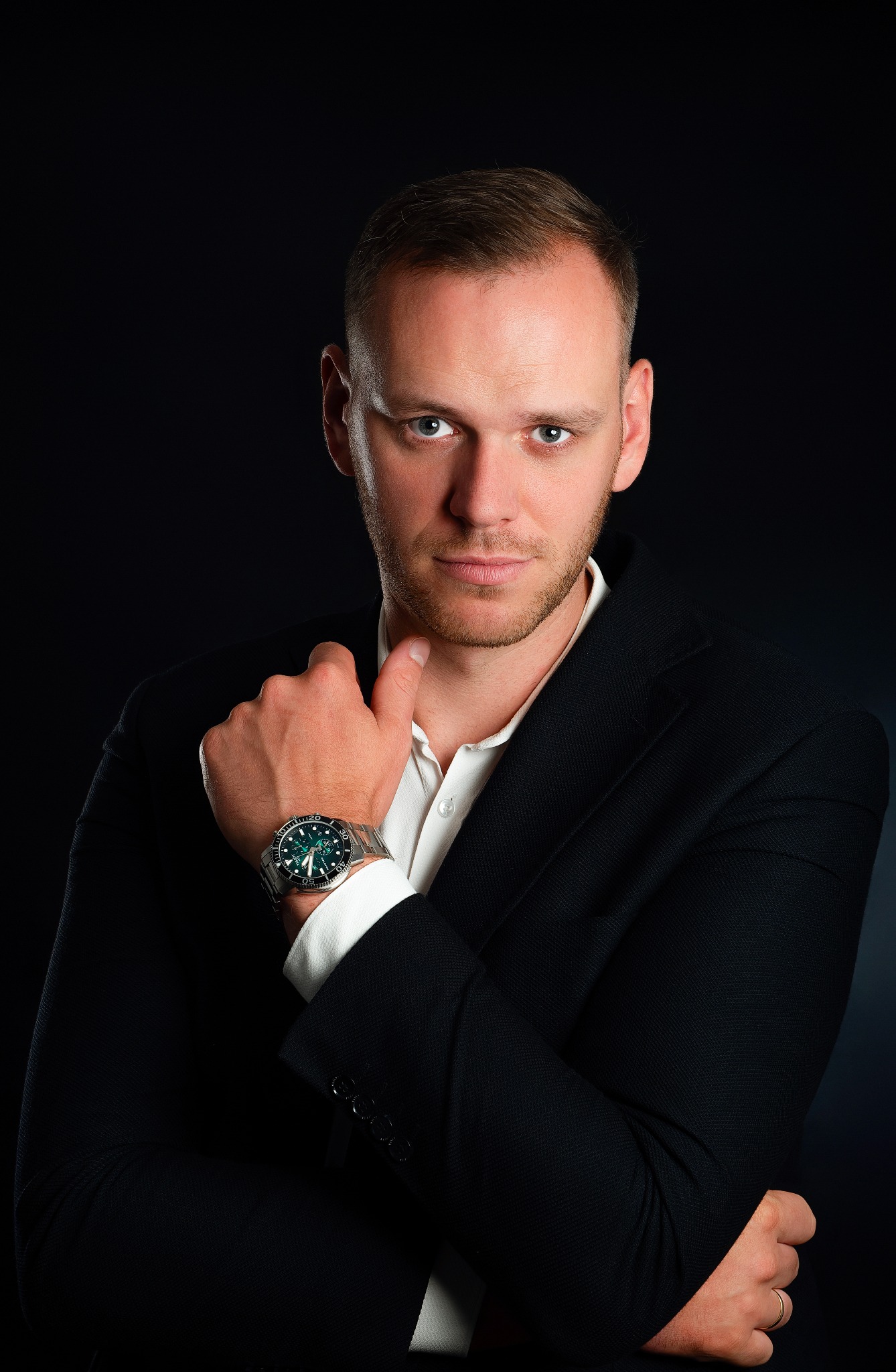 Portret mężczyzny w czarnej marynarce i białej koszuli, z zegarkiem na ręku, pozującego na ciemnym tle.