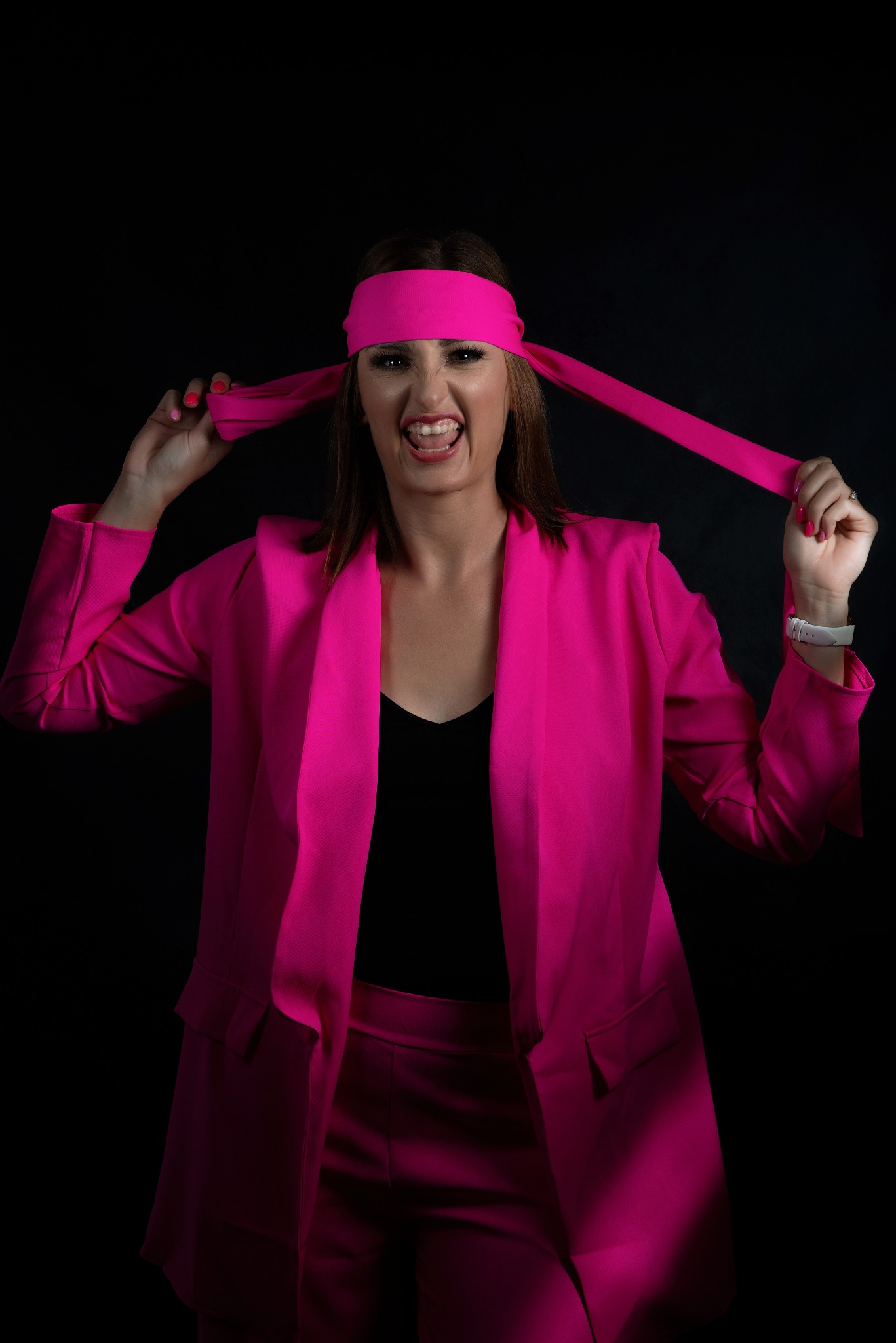 Kobieta w fuksjowym garniturze i opasce na głowie, z szerokim uśmiechem i wyrazistym makijażem na czarnym tle.