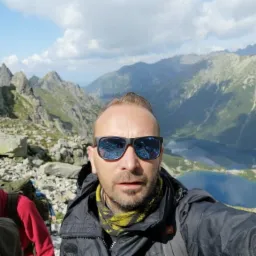 Selfie mężczyzny w okularach przeciwsłonecznych z widokiem na góry i jezioro w tle, z plecakiem i chustą na szyi, prawdopodobnie podczas wędrówki.