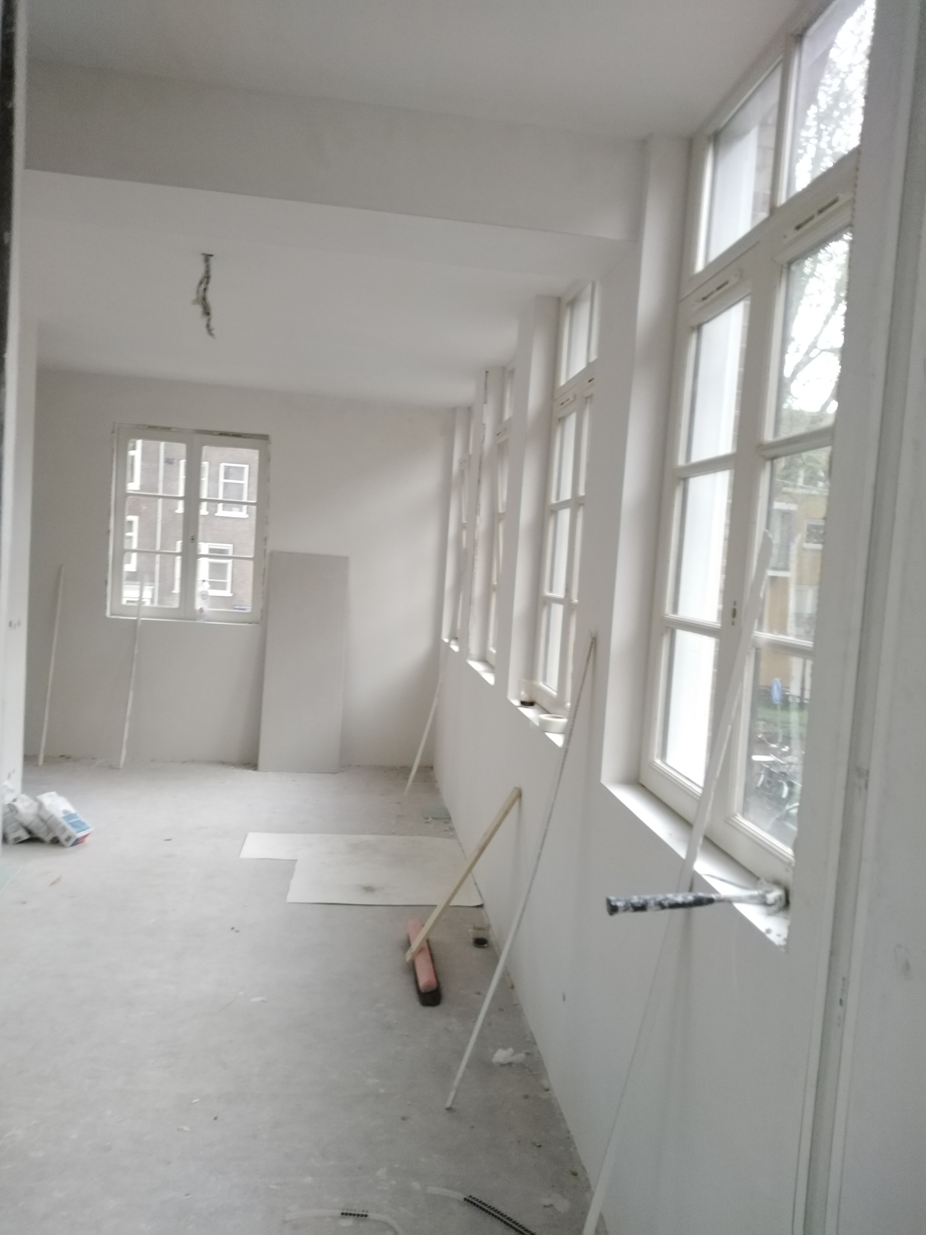 Wnętrze po nałożeniu białych tynków, widoczne okna z drewnianą stolarką, resztki materiałów budowlanych i narzędzia na podłodze, kabel zwisający z sufitu.
