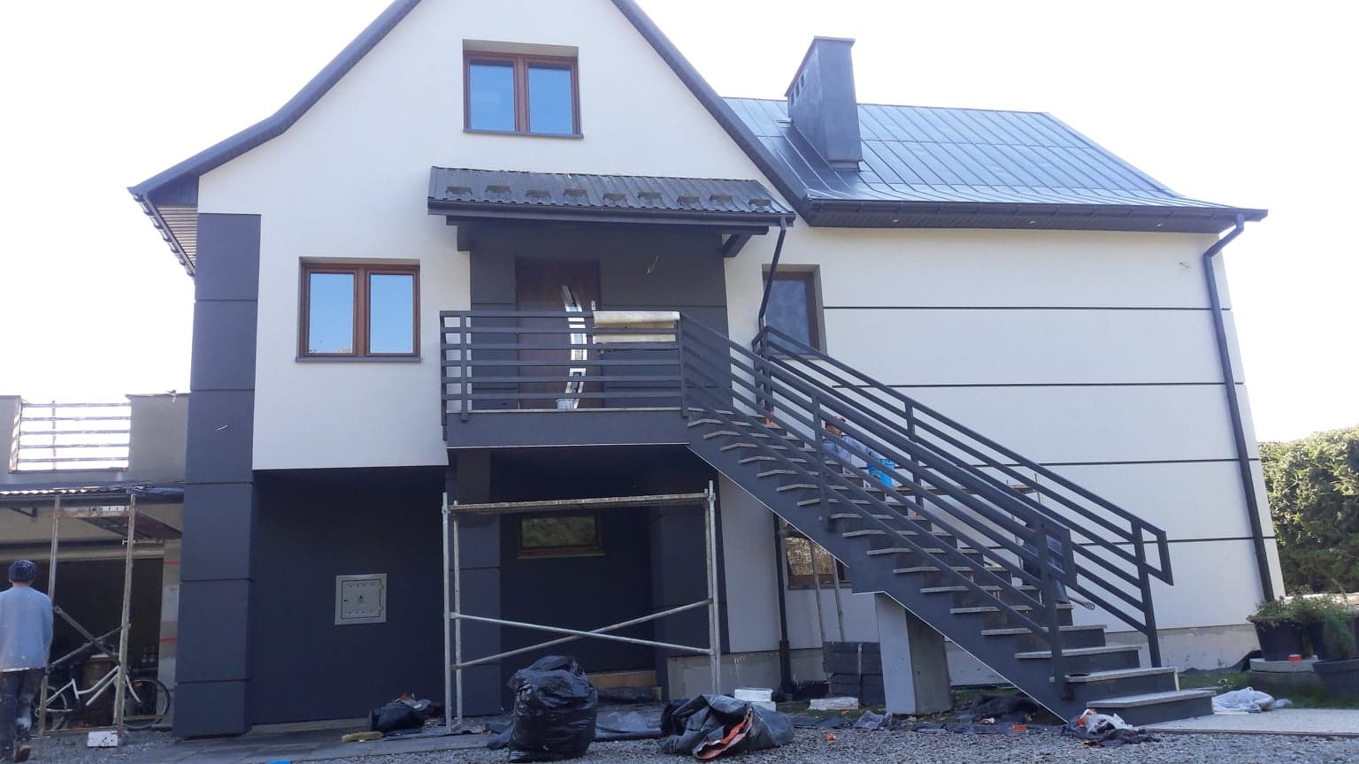 Nowoczesny dom z elewacją w kolorach bieli i grafitu, z metalowymi schodami zewnętrznymi i fragmentem rusztowania, widoczne ślady prac wykończeniowych wokół budynku.