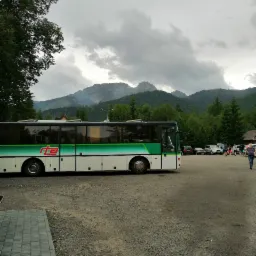 Autokar z zielonym pasem na tle górskiego krajobrazu i pochmurnego nieba, zaparkowany na żwirowym parkingu.