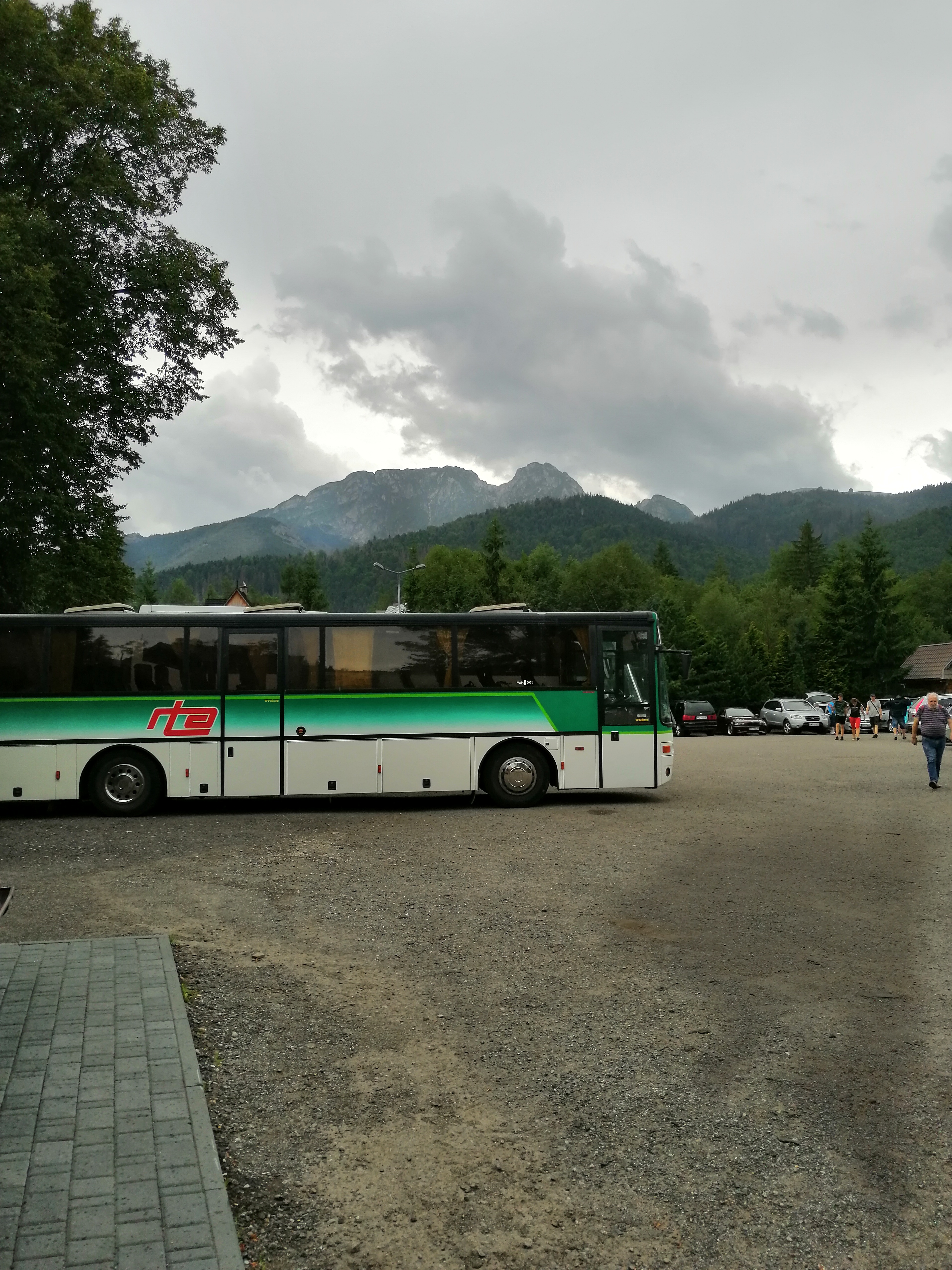 Autokar z zielonym pasem na tle górskiego krajobrazu i pochmurnego nieba, zaparkowany na żwirowym parkingu.