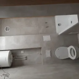 Wykończona łazienka z beżowymi płytkami, wiszącą muszlą klozetową, umywalką i podgrzewaczem wody Ariston, widoczne przyłącza i wentylacja.