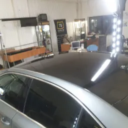 Szary samochód z przyciemnionymi szybami i dachem, sfotografowany w warsztacie z widocznym oświetleniem roboczym i banerem firmy Tomglass w tle.