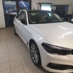 Białe BMW serii 5 z czarnym dachem, zaparkowane w warsztacie z jasnymi płytkami na podłodze i widocznym oświetleniem LED do detailingu.