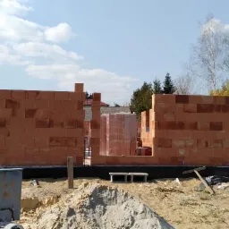 Wznoszenie ścian domu z czerwonej ceramiki, widoczna betonowa mieszarka, palety z materiałem budowlanym i fragment izolacji fundamentów.