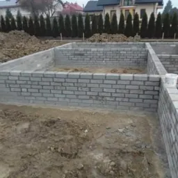 Wznoszenie ścian fundamentowych z bloczków betonowych, widoczne szalunki i przygotowane podłoże gruntowe, w tle dom w stanie surowym otwartym.