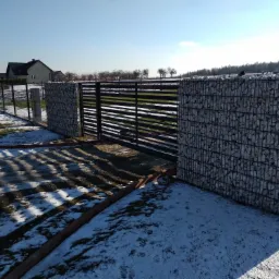 Nowoczesna brama wjazdowa z poziomymi przęsłami i ogrodzeniem z gabionów wypełnionych jasnymi kamieniami, z resztkami śniegu na trawniku w słoneczny dzień.