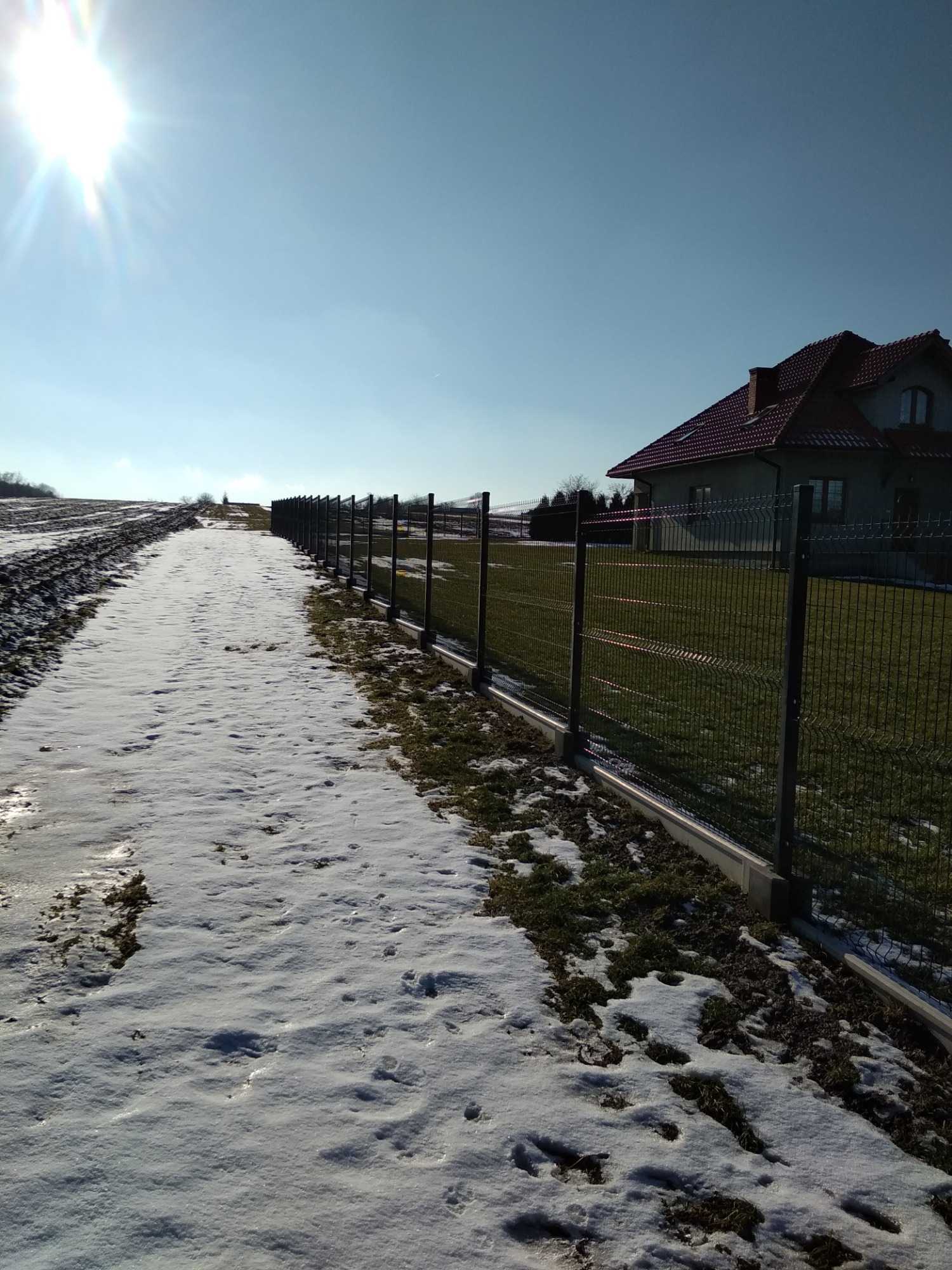 Długa perspektywa czarnego ogrodzenia panelowego na betonowej podmurówce, biegnącego wzdłuż zaśnieżonej drogi w kierunku domu z czerwoną dachówką, w słoneczny dzień.