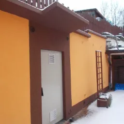 Budynek z pomarańczową elewacją i brązowymi detalami, widoczna brama garażowa, wentylacja w ścianie, drabinka drewniana i zasypany śniegiem teren.