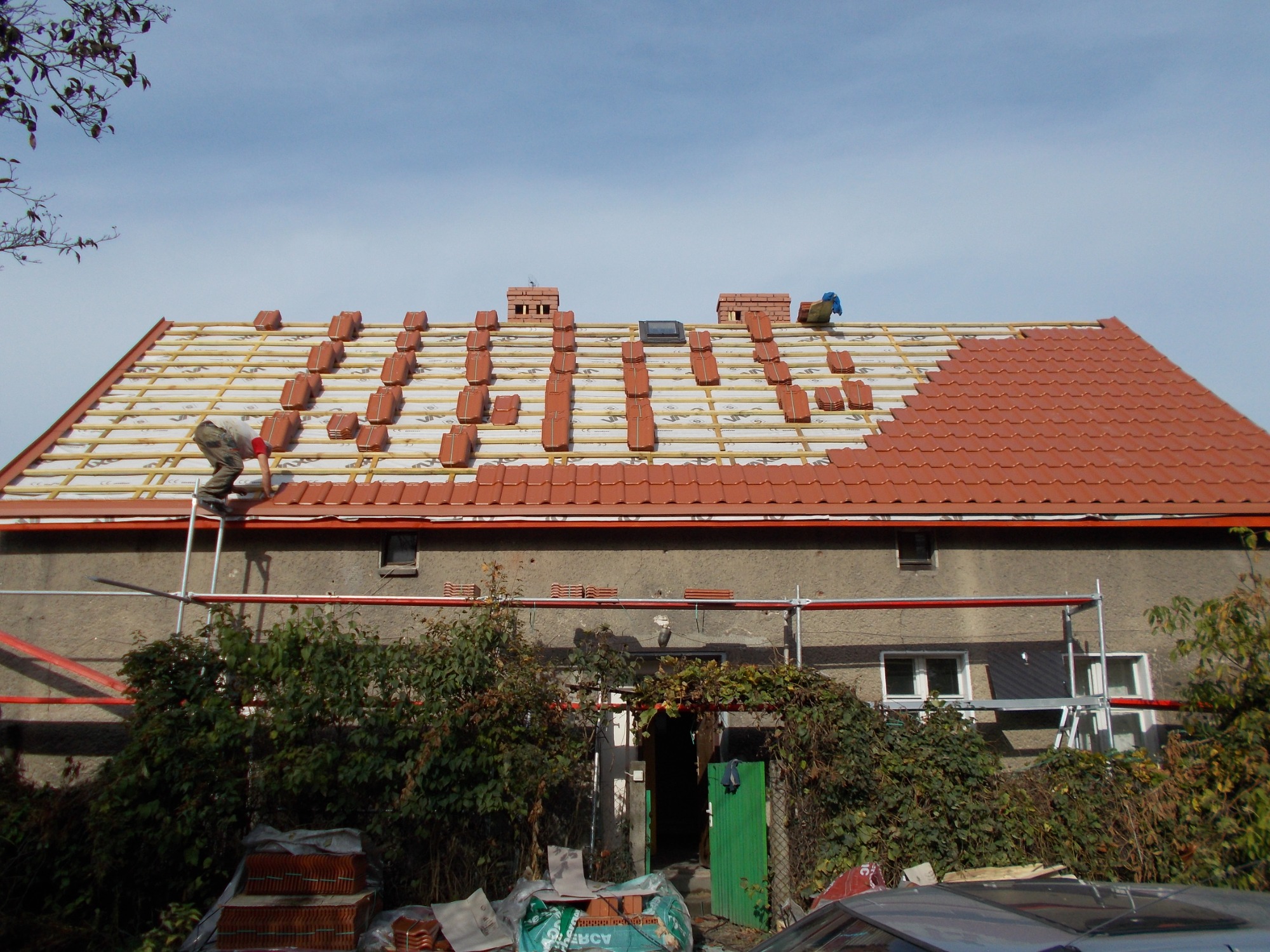 Montaż czerwonej dachówki na dachu domu jednorodzinnego; widoczne ułożone rzędy dachówek, częściowo odkryta konstrukcja dachu, kominy oraz pracownik na drabinie.