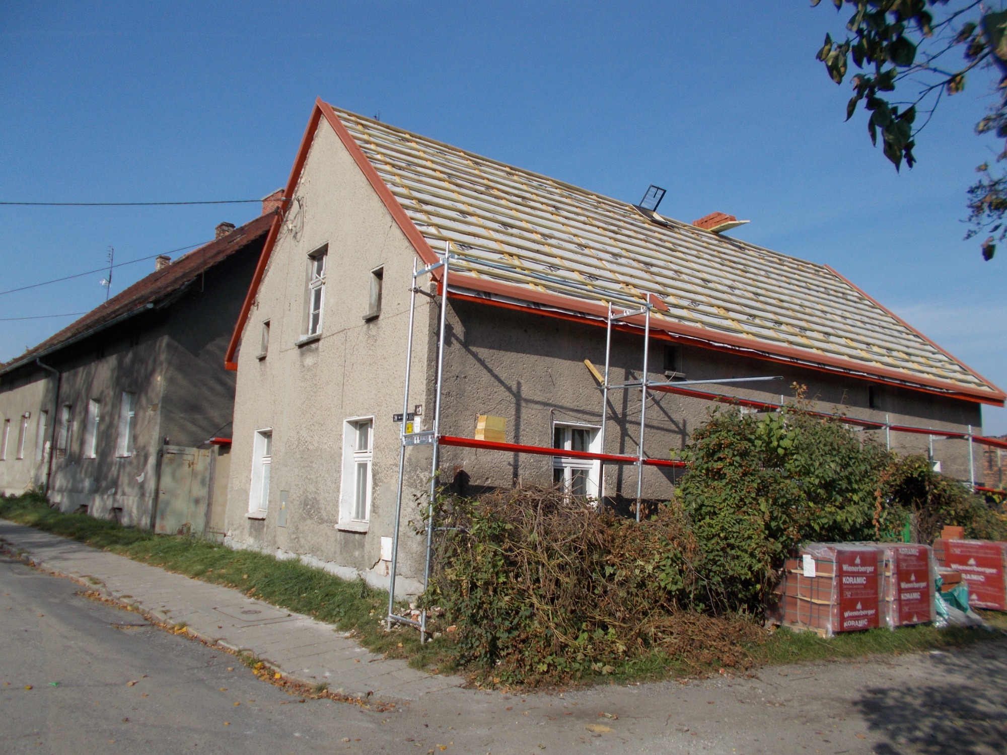 Dom z odkrytą konstrukcją dachu podczas remontu, widoczne rusztowania, palety z dachówkami Wienerberger Koramic oraz zarośla u podnóża ściany.