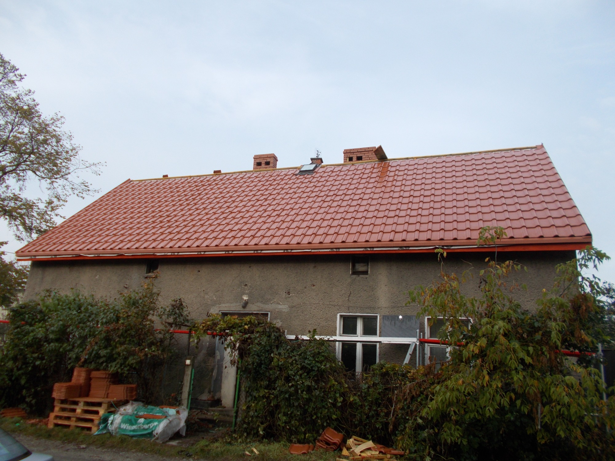Dom z nową, czerwoną dachówką ceramiczną, widoczne kominy i okno dachowe, częściowo zasłonięty przez roślinność, z paletą dachówek na pierwszym planie.