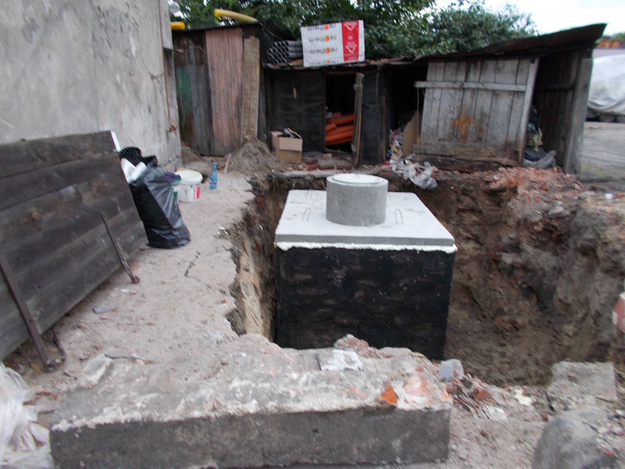 Betonowa studzienka kanalizacyjna w wykopie, zabezpieczona czarną izolacją, widok na zaniedbane podwórko z drewnianymi budynkami gospodarczymi i stertą gruzu.